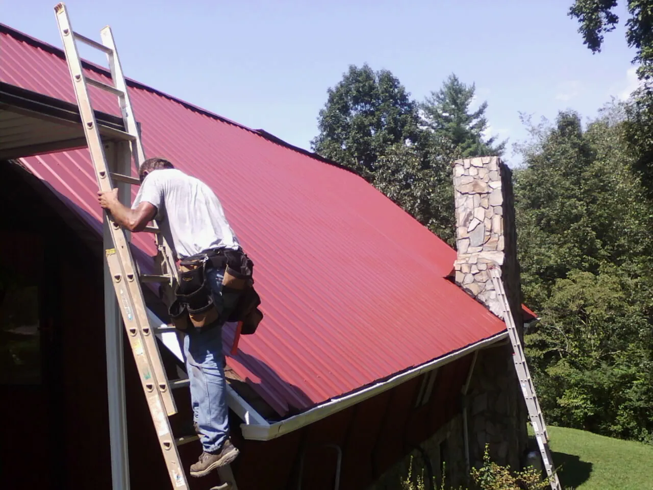 Roofer on a ladder beside a red metal roof and stone chimney — Soffit & Fascia Repair by Roofing Brothers in Darden